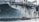 A large naval aircraft carrier with visible identification letters is stationed in the water. On the deck, various equipment and a helicopter are present. Below, a smaller patrol boat, manned by military personnel, navigates near a bright yellow buoy. The scene appears industrial and maritime.