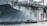A large naval aircraft carrier with visible identification letters is stationed in the water. On the deck, various equipment and a helicopter are present. Below, a smaller patrol boat, manned by military personnel, navigates near a bright yellow buoy. The scene appears industrial and maritime.