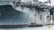 A large naval aircraft carrier with visible identification letters is stationed in the water. On the deck, various equipment and a helicopter are present. Below, a smaller patrol boat, manned by military personnel, navigates near a bright yellow buoy. The scene appears industrial and maritime.