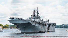 A large military aircraft carrier is stationed in a harbor, surrounded by water. The ship features a flat top deck and several antennae. Various smaller boats are visible in the surrounding area, including a small tugboat assisting the carrier. The sky is partly cloudy, and there are a few buildings and trees visible on the distant shore.