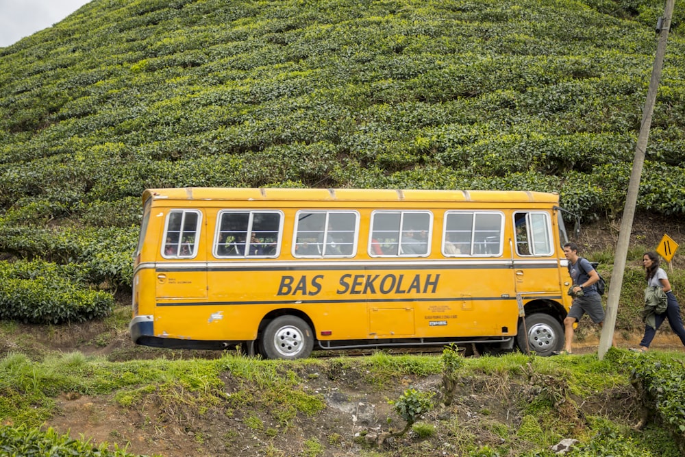 a yellow bus driving down a dirt road next to a lush green hillside