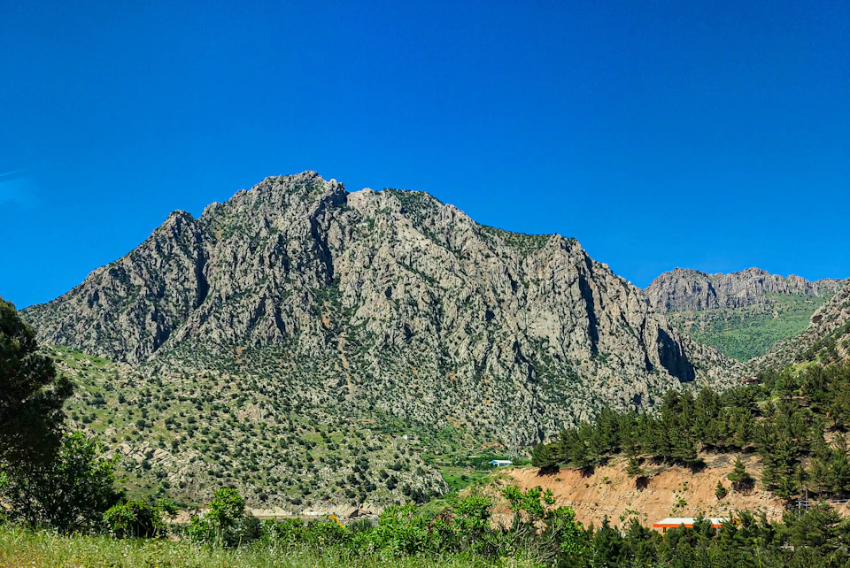 The image depicts a mountainous landscape with a large, jagged rock formation on the left side and a smaller, more rounded mountain range on the right side. The mountains are covered in lush green vegetation, creating a vibrant contrast against the clear blue sky above. The perspective of the photo is from ground level, looking up at the towering peaks, emphasizing their grandeur and scale.