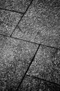 Close-up view of textured stone tiles arranged in a geometric pattern with visible seams. The surface is rough and speckled, creating an intricate design.