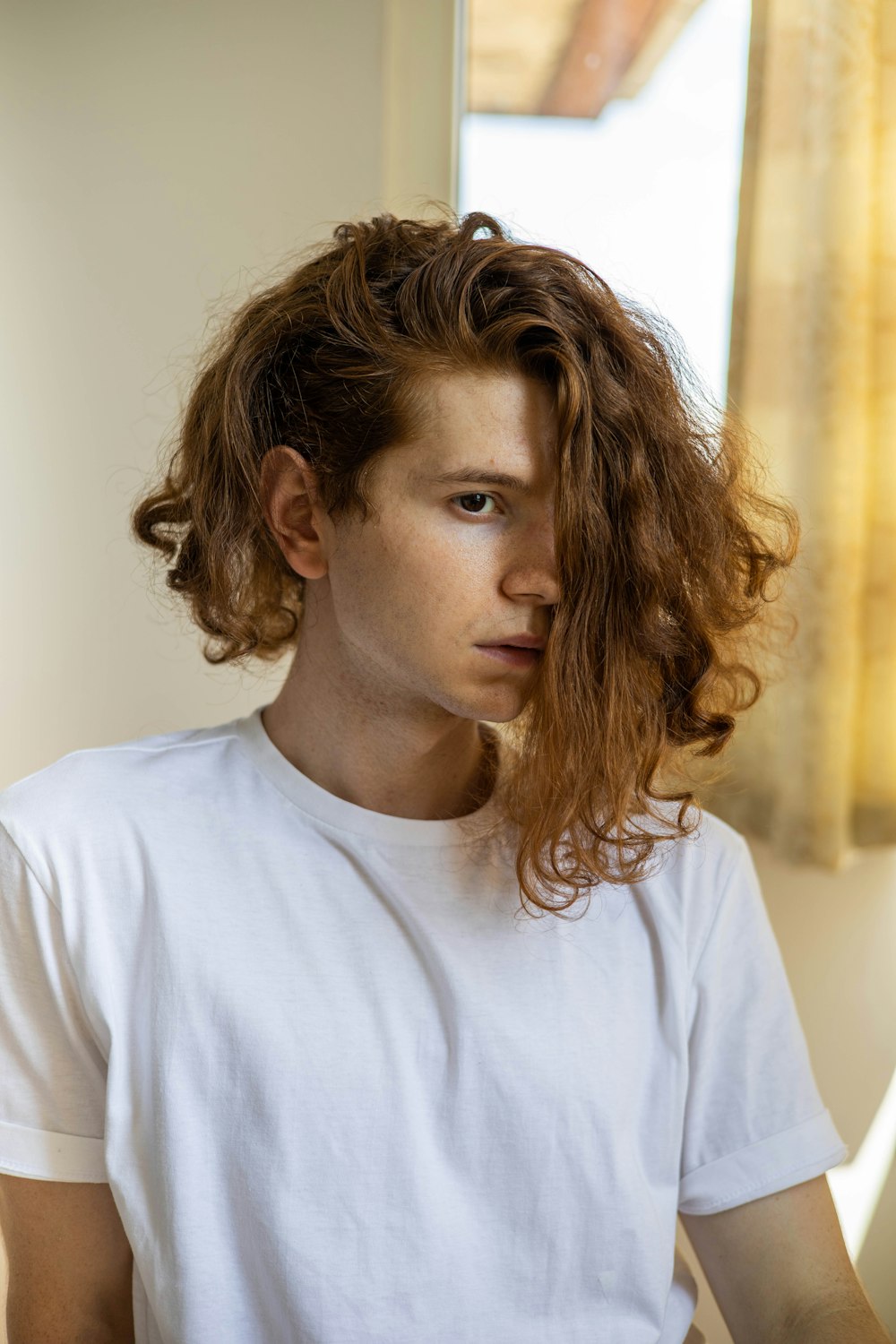 a young man with curly hair sitting in front of a window