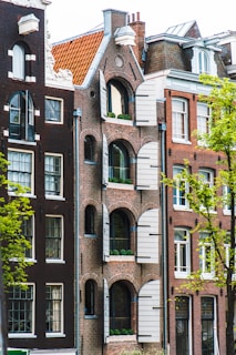 A row of traditional European-style buildings with intricate brickwork features arched windows with white shutters. The structures have a distinct architectural style, with elements such as steep, gabled roofs and decorative facade details. The scene includes a tree in the foreground with green leaves, suggesting a vibrant and lively atmosphere.
