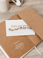 A minimalistic scene featuring a brown notebook partially open with a card placed inside. The card has decorative dried leaves printed on it and the phrase 'Write your story'. A beige ribbed candle and some decorative stones are placed on the wooden surface nearby.