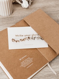 A minimalistic scene featuring a brown notebook partially open with a card placed inside. The card has decorative dried leaves printed on it and the phrase 'Write your story'. A beige ribbed candle and some decorative stones are placed on the wooden surface nearby.