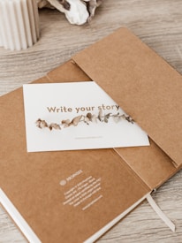 A minimalistic scene featuring a brown notebook partially open with a card placed inside. The card has decorative dried leaves printed on it and the phrase 'Write your story'. A beige ribbed candle and some decorative stones are placed on the wooden surface nearby.