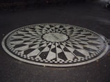 A circular black and white mosaic embedded in a paved surface. The word 'Imagine' is written in the center of the mosaic, surrounded by a patterned design reminiscent of a sunburst or starburst with alternating black and white segments. The area around the mosaic is dark and damp, suggesting recent rain. Fallen leaves are scattered on the ground.