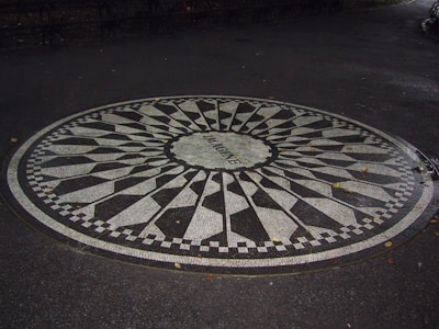 A circular black and white mosaic embedded in a paved surface. The word 'Imagine' is written in the center of the mosaic, surrounded by a patterned design reminiscent of a sunburst or starburst with alternating black and white segments. The area around the mosaic is dark and damp, suggesting recent rain. Fallen leaves are scattered on the ground.