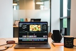 A laptop displaying video editing software on a desk, accompanied by a smartphone, a notepad with a pen, and a coffee mug with a motivational quote. The background shows a modern office environment with glass panels and some blurred details.