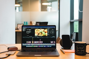 A laptop displaying video editing software on a desk, accompanied by a smartphone, a notepad with a pen, and a coffee mug with a motivational quote. The background shows a modern office environment with glass panels and some blurred details.