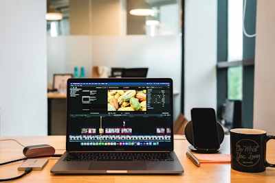 A laptop displaying video editing software on a desk, accompanied by a smartphone, a notepad with a pen, and a coffee mug with a motivational quote. The background shows a modern office environment with glass panels and some blurred details.