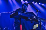 A musician plays a keyboard on stage, surrounded by a backdrop illuminated with blue and purple lighting. The scene includes a microphone stand and various cables attached to the equipment, creating a moody and atmospheric setting. The musician has a focused expression, contributing to the intense ambiance.