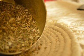 A close-up of a pile of dried herbs spilling from a container onto a textured, woven surface. The foreground is filled with the greenish-brown herbs, while the background is softly blurred.