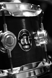 A close-up view of a drum with metallic components, showing a logo that indicates it is handcrafted and made in the USA. The image is in black and white, highlighting the texture and details of the drum's surface and hardware.