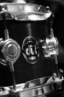 A close-up view of a drum with metallic components, showing a logo that indicates it is handcrafted and made in the USA. The image is in black and white, highlighting the texture and details of the drum's surface and hardware.