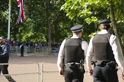 A few police officers stand in a park area with green trees in the background. They are wearing dark uniforms and hats, and seem to be monitoring a crowd of people gathered behind metal barricades. A Union Jack flag is visible on a pole to the left side.