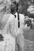 A black and white image of a couple dancing closely. The woman is wearing a lace wedding dress, and the man is dressed in a shirt with suspenders and a bow tie. They are holding hands, and the man kisses her cheek.