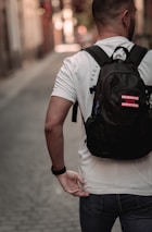 A person with short hair is walking down a narrow cobblestone street, wearing a white t-shirt and carrying a black backpack with a patch that says 'Bad decisions make good stories.' The street is lined with blurred buildings, indicating a shallow depth of field.