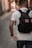 A person with short hair is walking down a narrow cobblestone street, wearing a white t-shirt and carrying a black backpack with a patch that says 'Bad decisions make good stories.' The street is lined with blurred buildings, indicating a shallow depth of field.