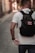 A person with short hair is walking down a narrow cobblestone street, wearing a white t-shirt and carrying a black backpack with a patch that says 'Bad decisions make good stories.' The street is lined with blurred buildings, indicating a shallow depth of field.