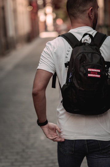 A person with short hair is walking down a narrow cobblestone street, wearing a white t-shirt and carrying a black backpack with a patch that says 'Bad decisions make good stories.' The street is lined with blurred buildings, indicating a shallow depth of field.