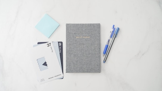 A neatly arranged flat lay of stationery, featuring a grey textured bullet journal at the center. On the left, there are two pens, one blue and one black, positioned parallel. To the right, there are a series of swatch cards displaying colors labeled 'Deep Fog' and 'Navy Blue'. A light blue sticky note is partially visible to the top left.