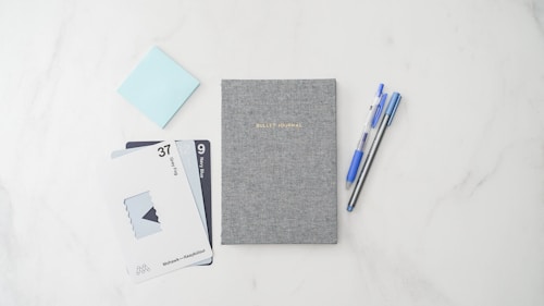 A neatly arranged flat lay of stationery, featuring a grey textured bullet journal at the center. On the left, there are two pens, one blue and one black, positioned parallel. To the right, there are a series of swatch cards displaying colors labeled 'Deep Fog' and 'Navy Blue'. A light blue sticky note is partially visible to the top left.