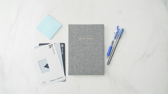 A neatly arranged flat lay of stationery, featuring a grey textured bullet journal at the center. On the left, there are two pens, one blue and one black, positioned parallel. To the right, there are a series of swatch cards displaying colors labeled 'Deep Fog' and 'Navy Blue'. A light blue sticky note is partially visible to the top left.
