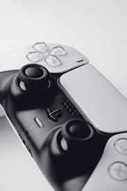 A close-up view of a modern gaming controller featuring a sleek black and white design. The image highlights the analog sticks and various buttons, including directional buttons and a central logo. The controller is set against a soft, neutral background, focusing attention on its details.