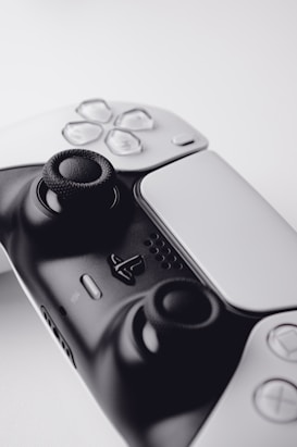 A close-up view of a modern gaming controller featuring a sleek black and white design. The image highlights the analog sticks and various buttons, including directional buttons and a central logo. The controller is set against a soft, neutral background, focusing attention on its details.