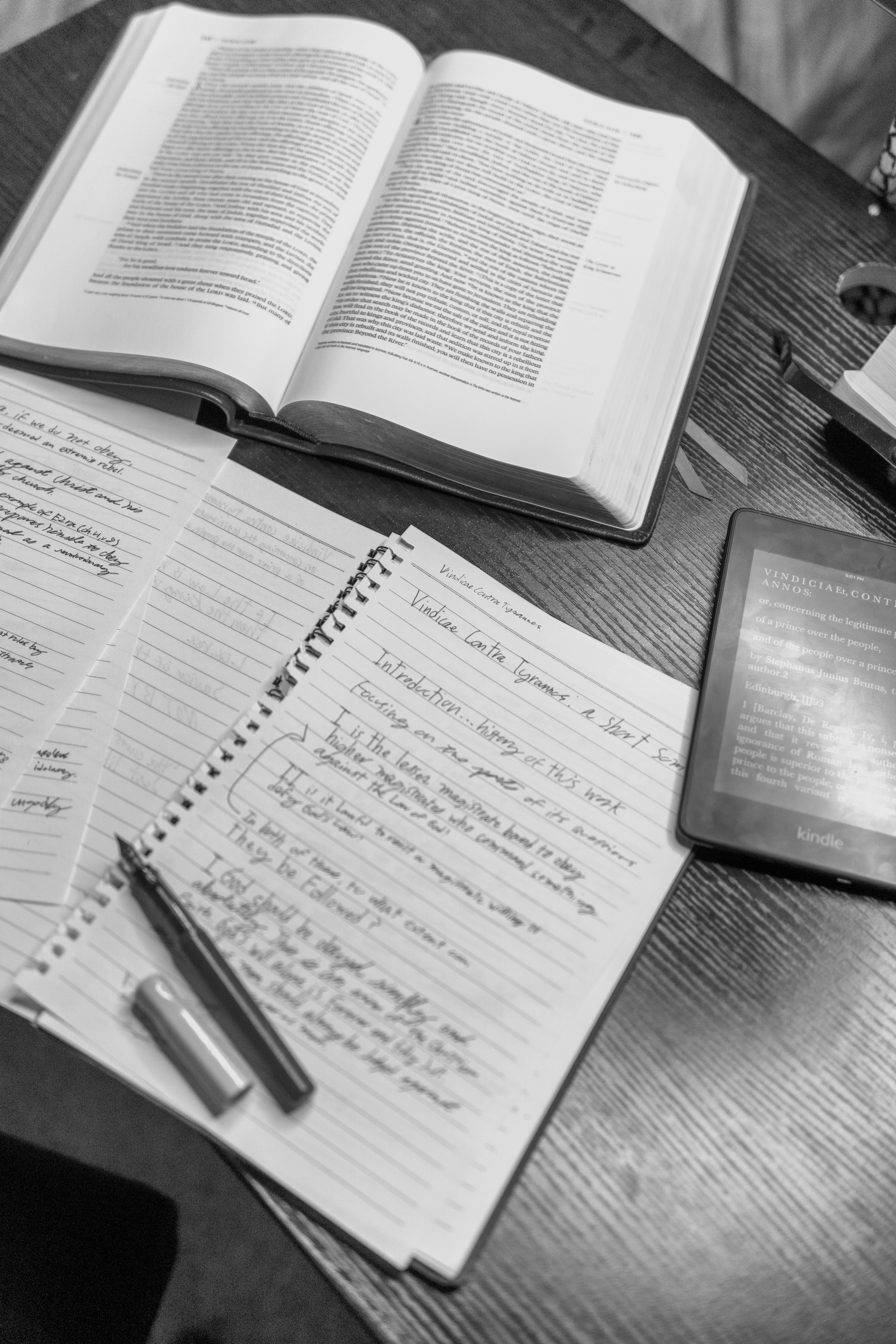 Open book, notebook, and e-reader on a wooden desk.
