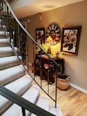 Cozy interior hallway with a carpeted staircase featuring decorative black railings. A round wall clock with bold numerals and stylish artwork adorn the light-colored walls. There is a wooden side table with a lit lamp, books, and a basket underneath. The hardwood flooring complements the warm, inviting atmosphere.