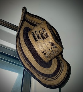 A large woven straw hat with intricate patterns hangs on a wall corner near a window. The hat features alternating dark and light bands, showcasing detailed craftsmanship.