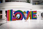 Multicolored, striped letters spell out the word LOVE inside a modern, spacious interior setting. A man in a striped shirt and cap photographs the installation, while two children play and interact with the letters.