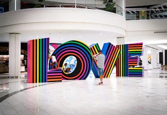 Multicolored, striped letters spell out the word LOVE inside a modern, spacious interior setting. A man in a striped shirt and cap photographs the installation, while two children play and interact with the letters.