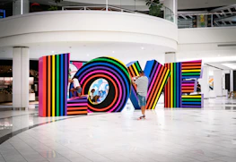 Multicolored, striped letters spell out the word LOVE inside a modern, spacious interior setting. A man in a striped shirt and cap photographs the installation, while two children play and interact with the letters.