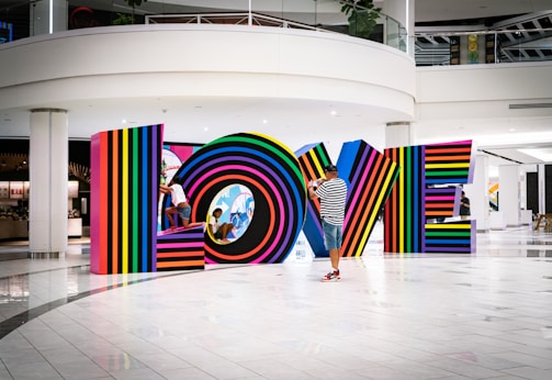 Multicolored, striped letters spell out the word LOVE inside a modern, spacious interior setting. A man in a striped shirt and cap photographs the installation, while two children play and interact with the letters.