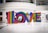 Multicolored, striped letters spell out the word LOVE inside a modern, spacious interior setting. A man in a striped shirt and cap photographs the installation, while two children play and interact with the letters.