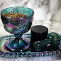 An iridescent glass goblet filled with a herbal substance is placed on a matching tray. Beside it, there is a black container and a green glass pipe. The background is a marble surface, adding an elegant feel to the setup.