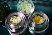 Several small glass jars contain different types of crystalline or granular substances, placed on a reflective surface. The contents vary in color and texture, with one jar holding a pale yellow, crumbly substance and another containing a darker amber, granular material.