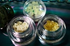 Several small glass jars contain different types of crystalline or granular substances, placed on a reflective surface. The contents vary in color and texture, with one jar holding a pale yellow, crumbly substance and another containing a darker amber, granular material.