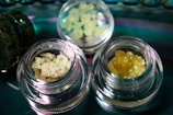 Several small glass jars contain different types of crystalline or granular substances, placed on a reflective surface. The contents vary in color and texture, with one jar holding a pale yellow, crumbly substance and another containing a darker amber, granular material.