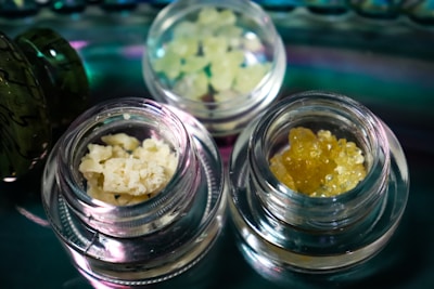 Several small glass jars contain different types of crystalline or granular substances, placed on a reflective surface. The contents vary in color and texture, with one jar holding a pale yellow, crumbly substance and another containing a darker amber, granular material.