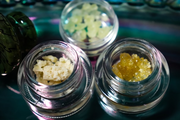 Several small glass jars contain different types of crystalline or granular substances, placed on a reflective surface. The contents vary in color and texture, with one jar holding a pale yellow, crumbly substance and another containing a darker amber, granular material.