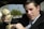 A person wearing a suit is sitting in a vehicle and holding an old-style beige telephone receiver. The background shows a blurred outdoor scene with greenery.