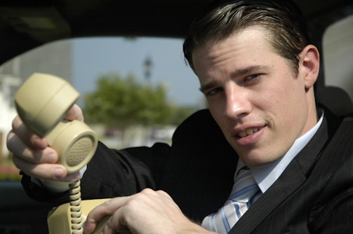 A person wearing a suit is sitting in a vehicle and holding an old-style beige telephone receiver. The background shows a blurred outdoor scene with greenery.
