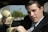 A person wearing a suit is sitting in a vehicle and holding an old-style beige telephone receiver. The background shows a blurred outdoor scene with greenery.
