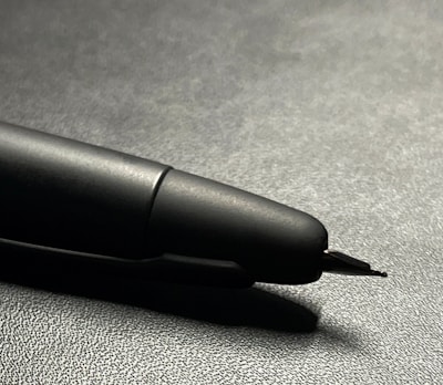 Close-up of the pen's tip with ink flowing in a deep black shade, set against a minimalist ivory background.